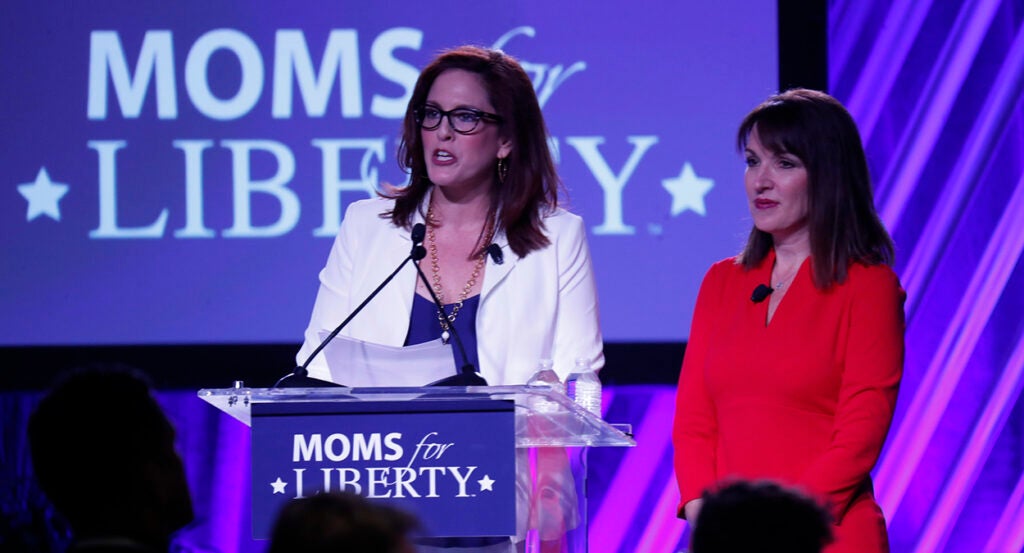 Tiffany Justice, in a blue dress and a white blazer, speaks in front of a podium reading "Moms for Liberty," as Tina Descovich, in a red dress, looks on.