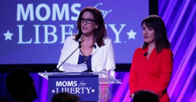 Tiffany Justice, in a blue dress and a white blazer, speaks in front of a podium reading "Moms for Liberty," as Tina Descovich, in a red dress, looks on.