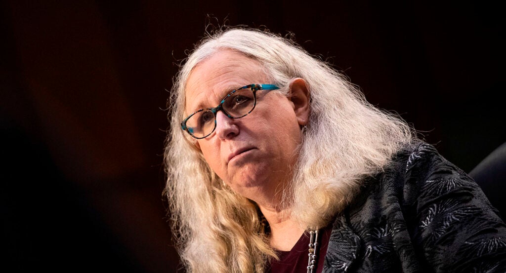 Rachel Levine in a dark blouse with masculine features, long blond hair, and glasses