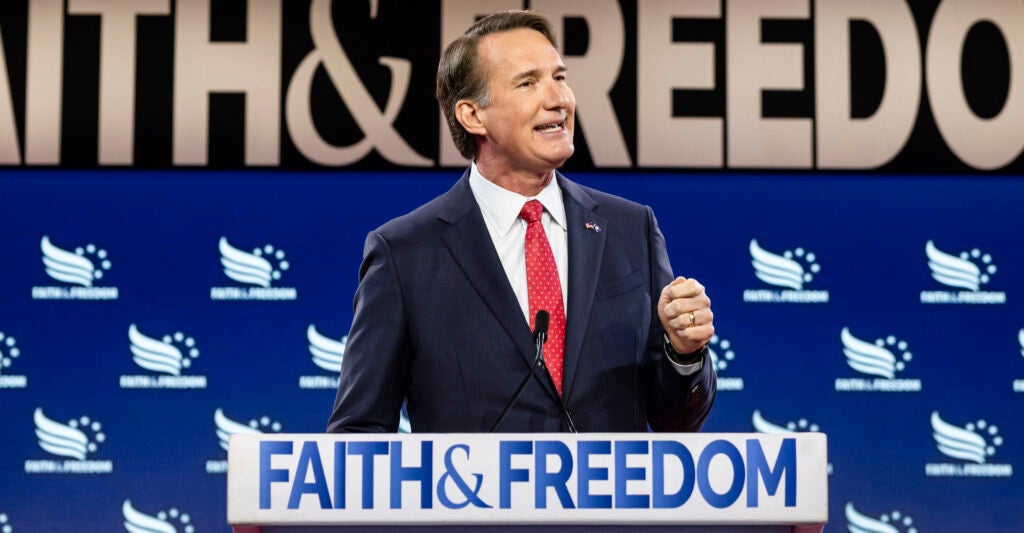 Governor Glenn Youngkin in a suit at a podium that says faith and freedom on the front, in front of a blue background with faith and freedom on it