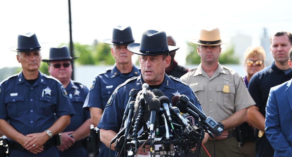 A sheriff speaks at a press conference with multiple microphones surrounded by other officers in uniform.