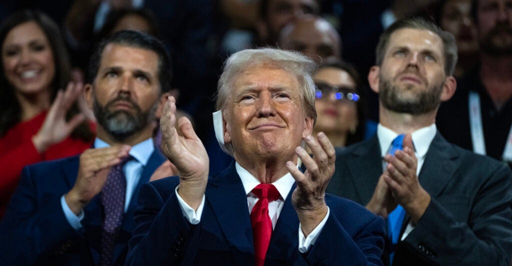 Former President Donald Trump, in a suit and clapping, pictured with his sons Don Jr. and Eric