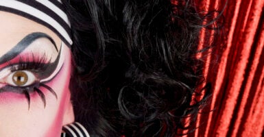 Half of a drag queen's face looks at the camera with a red curtain background.