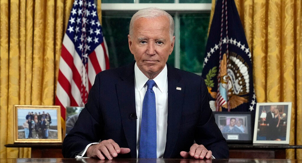 President Joe Biden in a black suit with a blue tie in the Oval Office