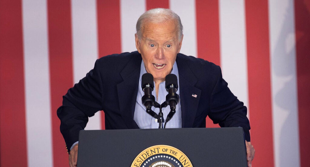 Joe Biden in a blue suit leans forward on a podium with the presidential seal