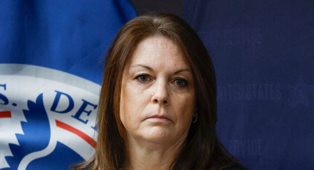 Secret Service Director Kimberly Cheatle stares into the camera in front of a seal for the Department of Homeland Security.