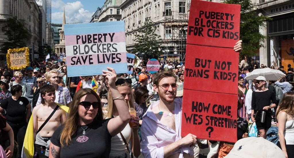 Protesters march with signs reading "Puberty Blockers Save Lives" and "Puberty Blockers for Cis Kids but Not Trans Kids"