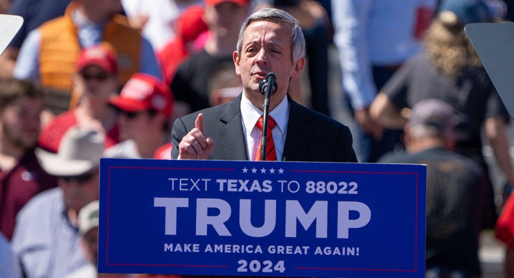 Robert Jeffress in a suit stands behind a podium reading "TRUMP Make America Great Again!"