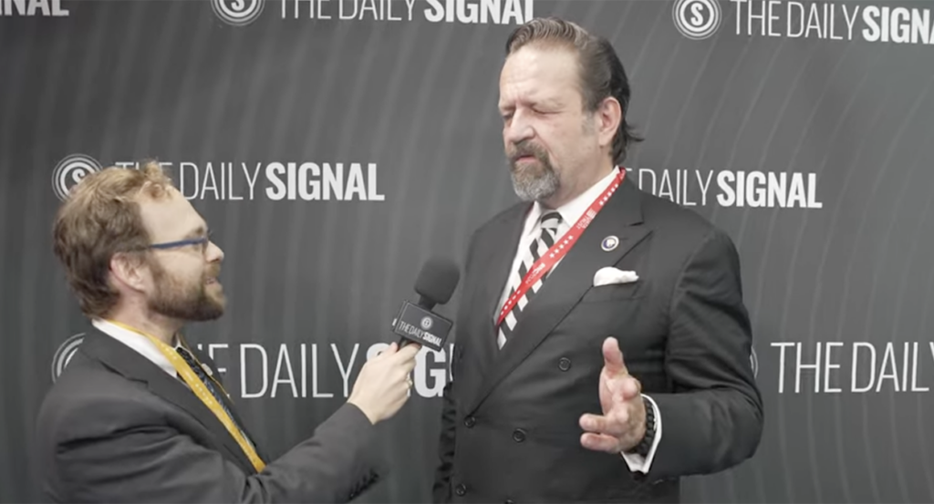 Tyler O'Neil in a grey suit holds the microphone for Seb Gorka in a black suit, gesturing.