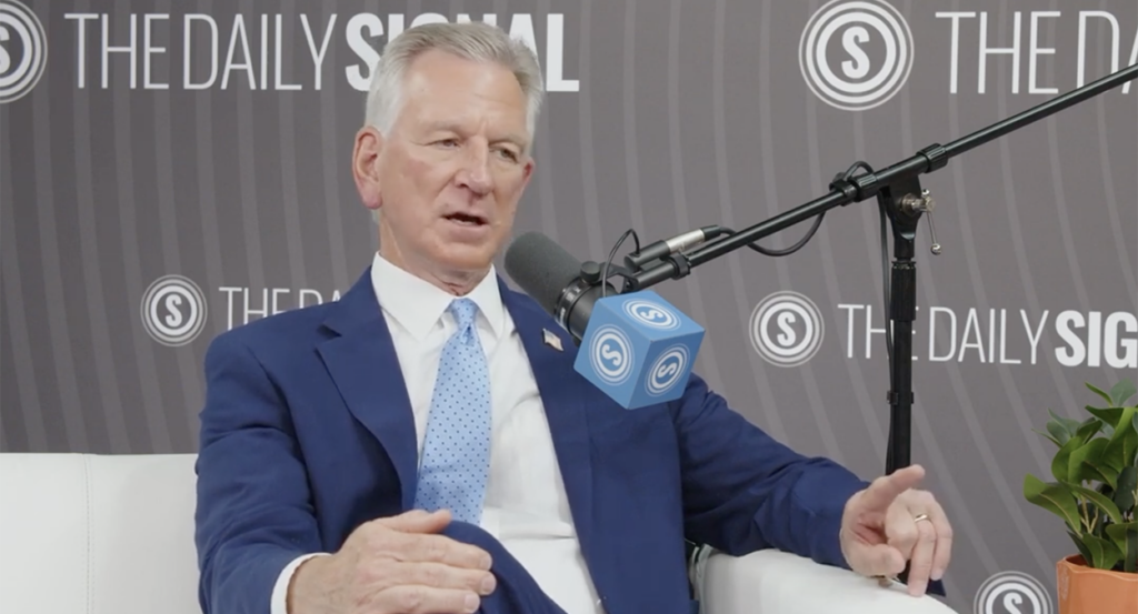 Tommy Tuberville in a blue suit sits in front of The Daily Signal