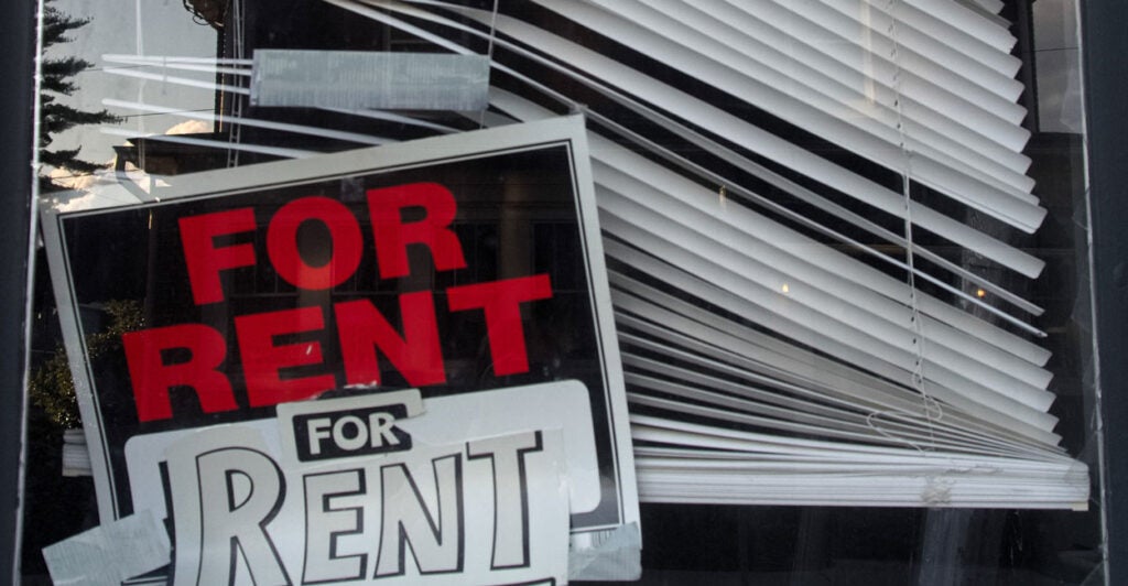 A crooked "For Rent" sign in the corner of a window with dilapidated blinds.