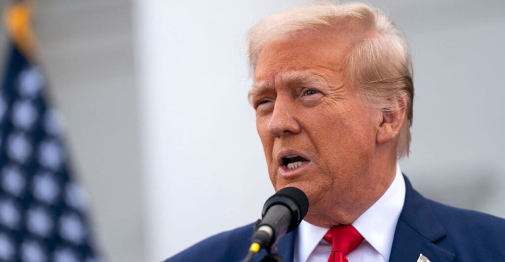 Donald Trump in a blue suit and a red tie at a microphone with an American flag in the background