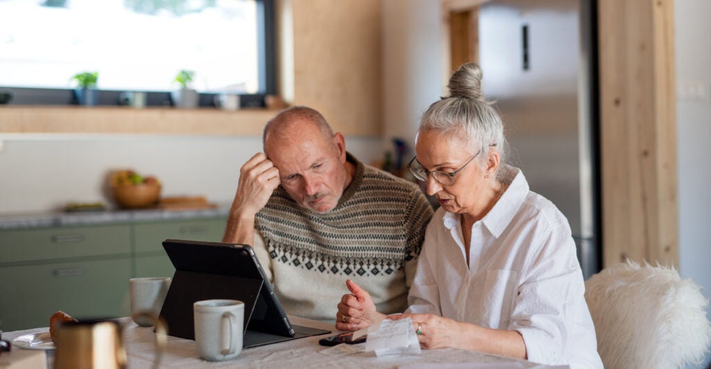 Senior couple sitting at the kitchen table with croissant and cups of tea, looking at digital tablet and recalculating their expenses.