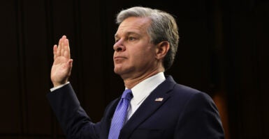 FBI Director Christopher Wray holds his hand up in a black suit.