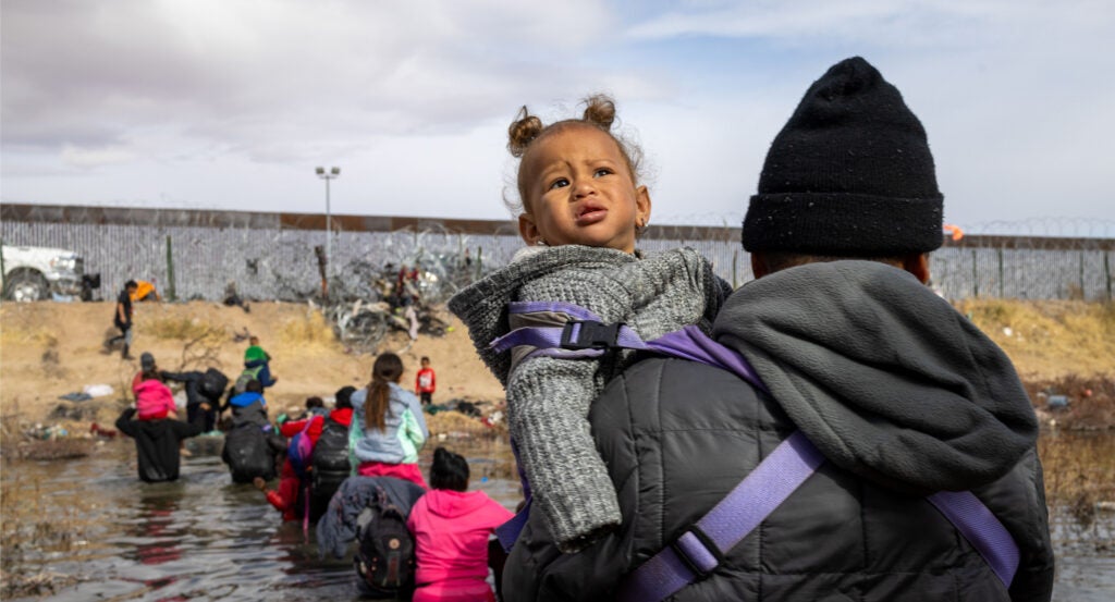 Migrant in black hoodie holds child in gray hoodie while crossing Rio Grande river with U.S. border wall in background.