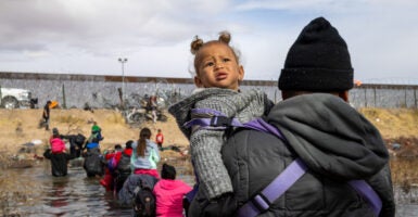 Migrant in black hoodie holds child in gray hoodie while crossing Rio Grande river with U.S. border wall in background.