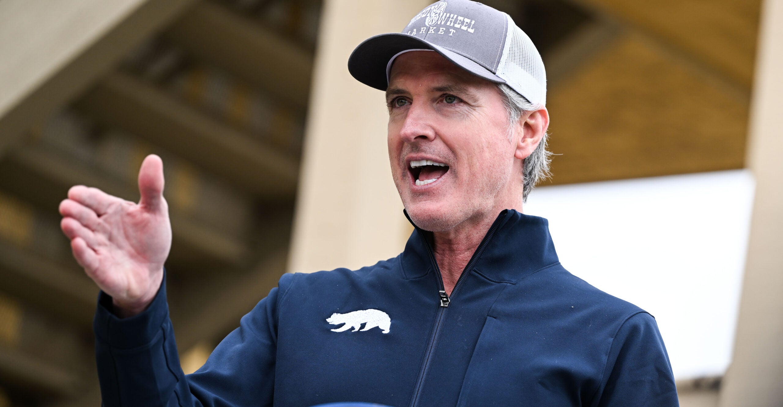 Gavin Newsom wears a ball cap, speaking with his hands.