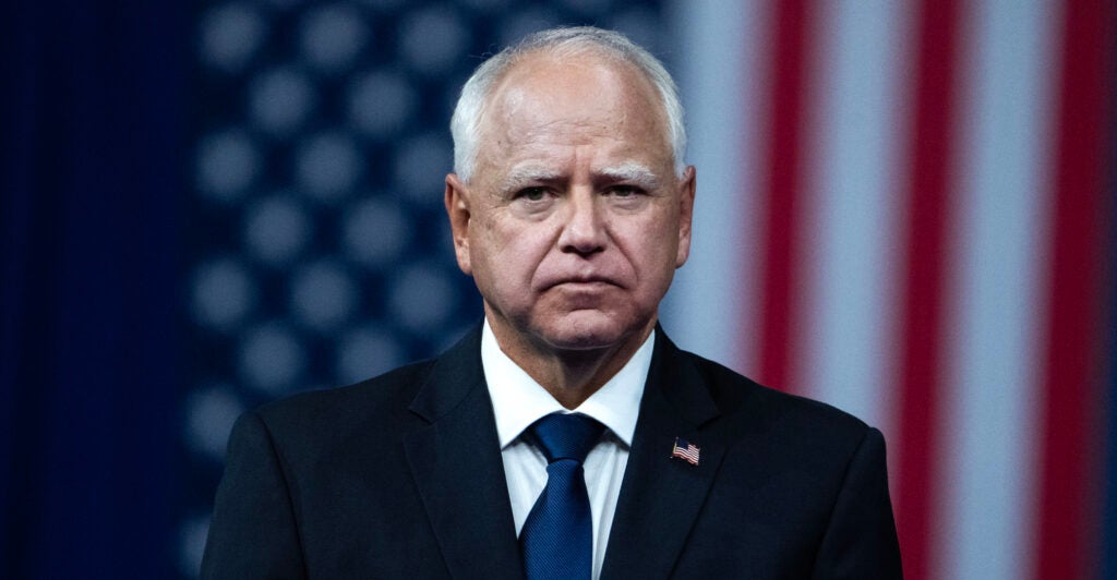 Walz stares grimly at the camera with a U.S. flag hanging behind him.