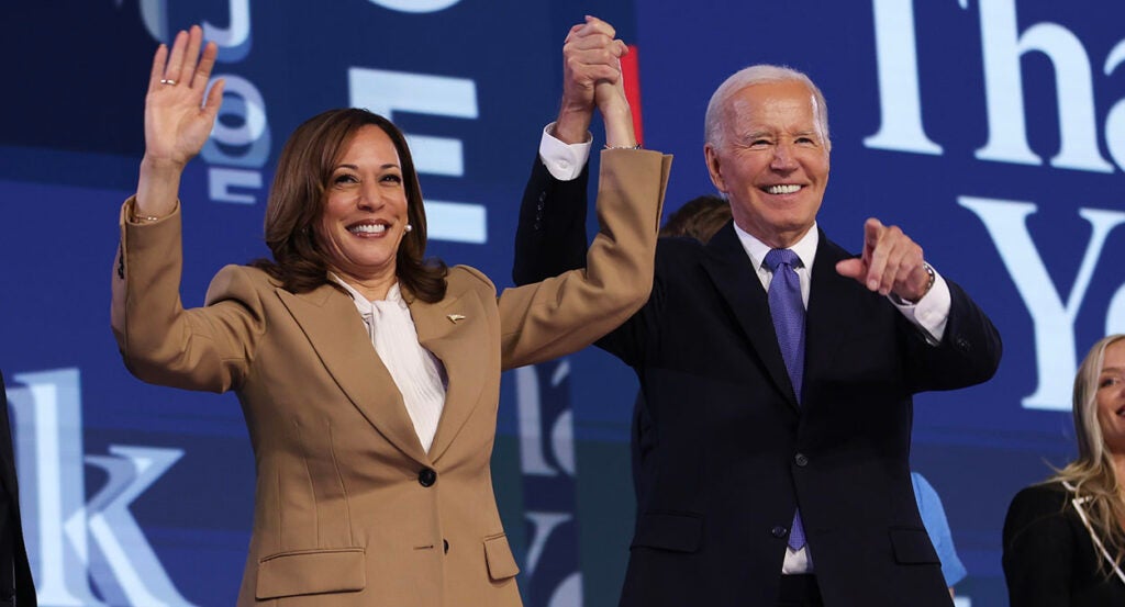 Kamala Harris in a tan coat holds hands with Joe Biden in a black suit