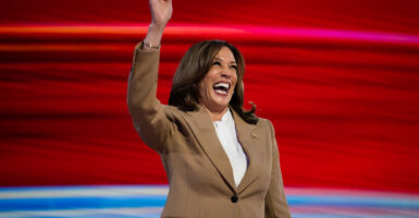 Kamala Harris on stage with a red background in a light brown suit smiling and waving