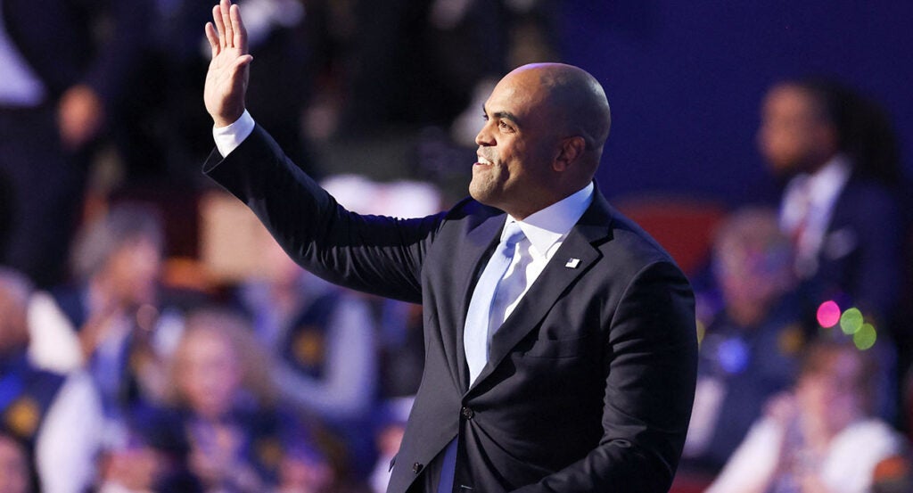 Colin Allred waves while wearing a black suit and a light blue tie