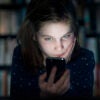 a young girl in a dark room in front of a shelf full of books looking at a smart phone with her face glowing from the phone's light