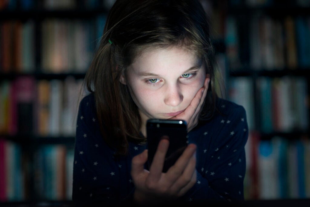 a young girl in a dark room in front of a shelf full of books looking at a smart phone with her face glowing from the phone's light