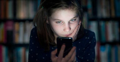 a young girl in a dark room in front of a shelf full of books looking at a smart phone with her face glowing from the phone's light