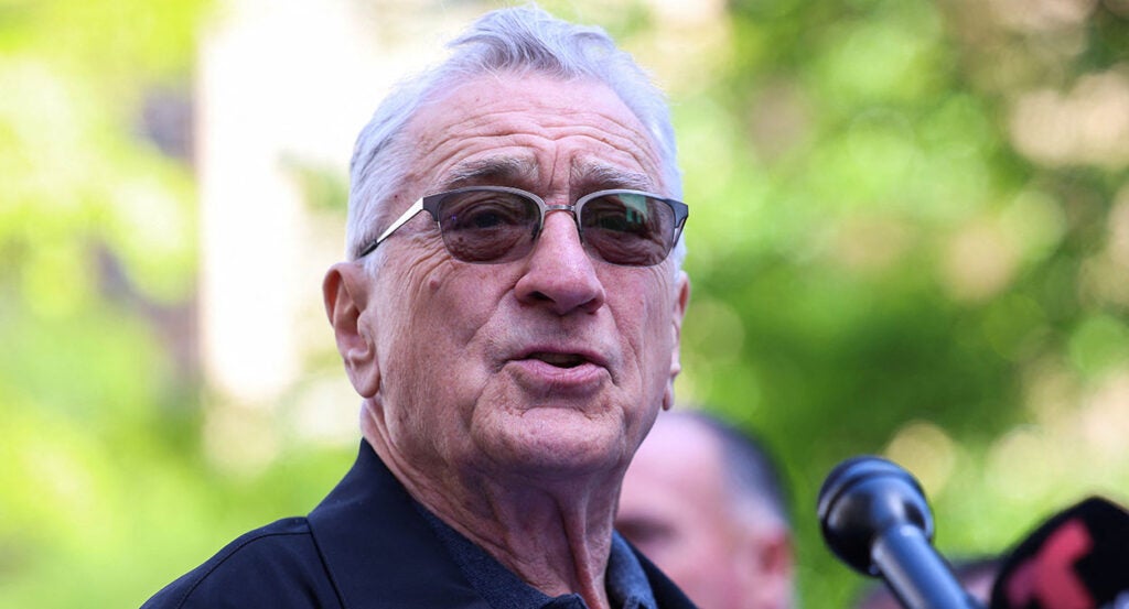 Robert DeNiro wears sunglasses and stands at a microphone