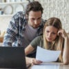 Unhappy young couple look at bill at kitchen table with laptop