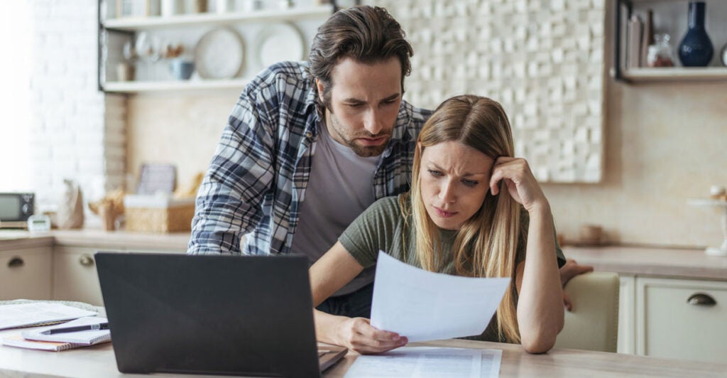 Unhappy young couple look at bill at kitchen table with laptop