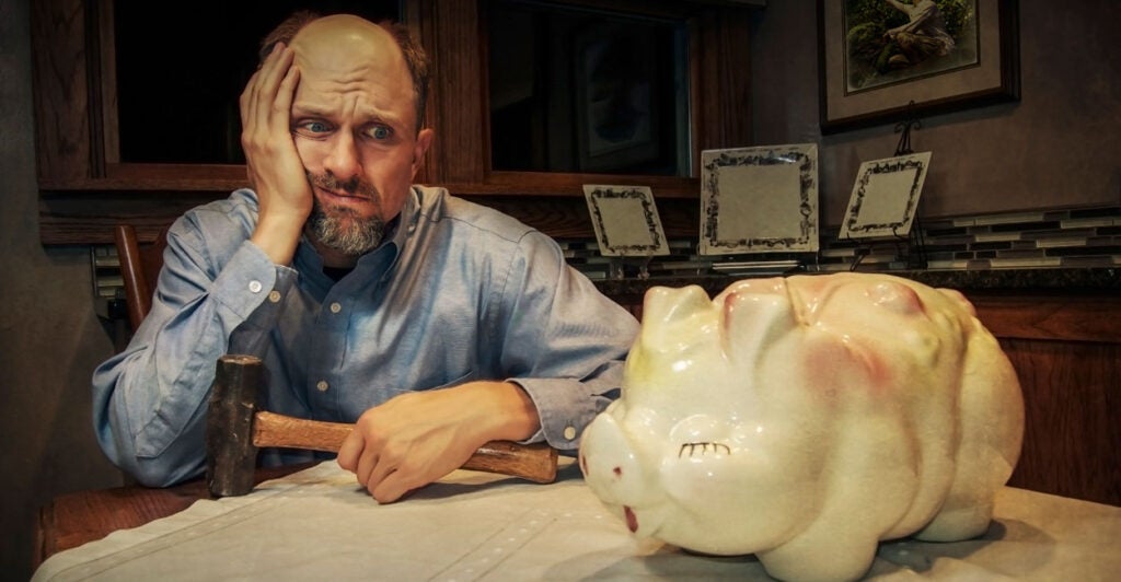 man with a hammer at a kitchen table debating smashing piggy bank