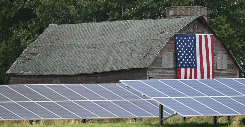 Solar panels on the ground in front of an old barn with an American flag hanging on the front of it.