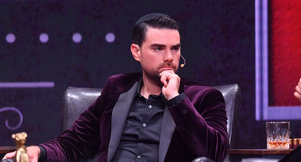 Ben Shapiro stares intently while stroking his beard in a black suit