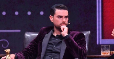 Ben Shapiro stares intently while stroking his beard in a black suit