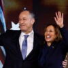 Doug Emhoff in a black suit hugs Kamala Harris in a black suit, as both stand in front of American flags.