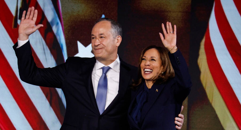 Doug Emhoff in a black suit hugs Kamala Harris in a black suit, as both stand in front of American flags.