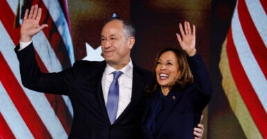 Doug Emhoff in a black suit hugs Kamala Harris in a black suit, as both stand in front of American flags.