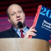 Jared Polis, in a blue suit, holds the Mandate for Leadership Project 2025 book