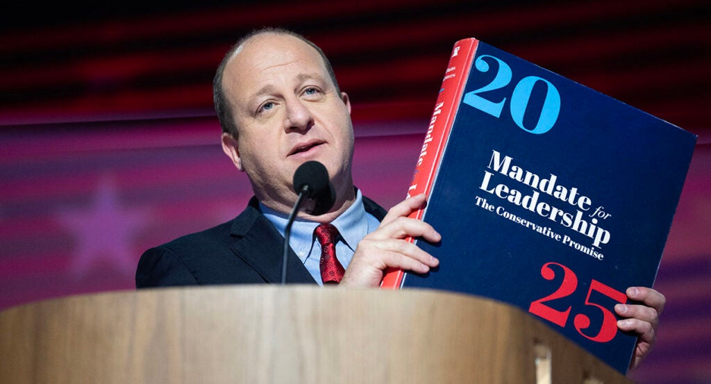 Jared Polis, in a blue suit, holds the Mandate for Leadership Project 2025 book