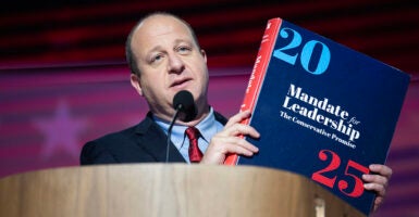 Jared Polis, in a blue suit, holds the Mandate for Leadership Project 2025 book