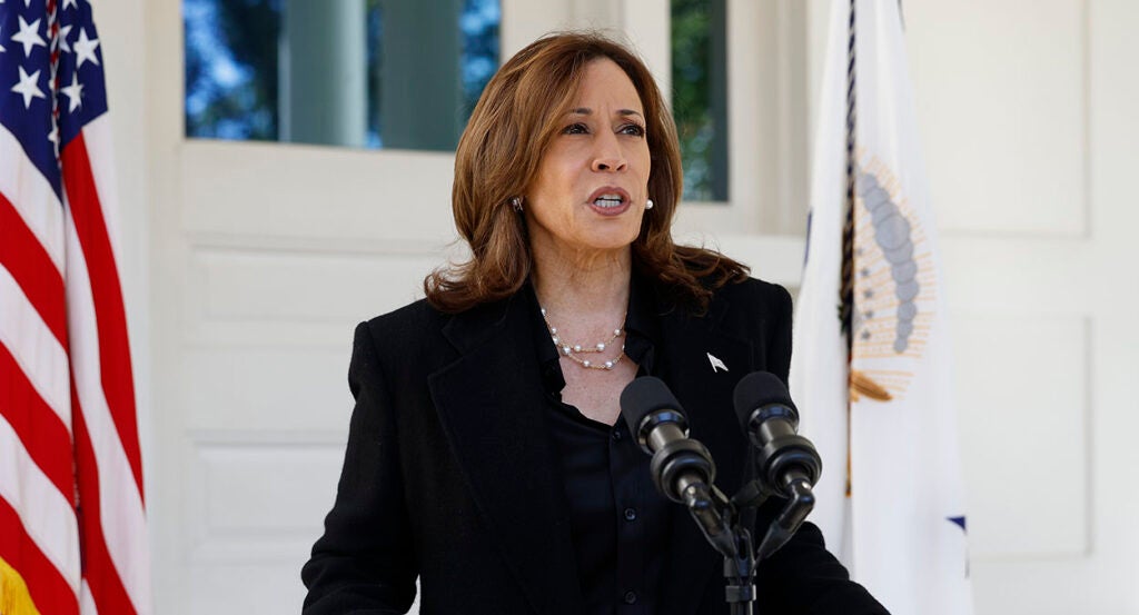 Kamala Harris in a black suit stands behind a microphone in front of an American flag.