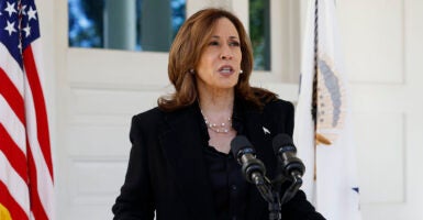 Kamala Harris in a black suit stands behind a microphone in front of an American flag.