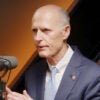 Rick Scott in a grey suit speaks in front of a microphone