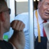 Donald Trump in a tie and McDonald's apron working a McDonald's drive-thru window, handing a bag of food to a customer