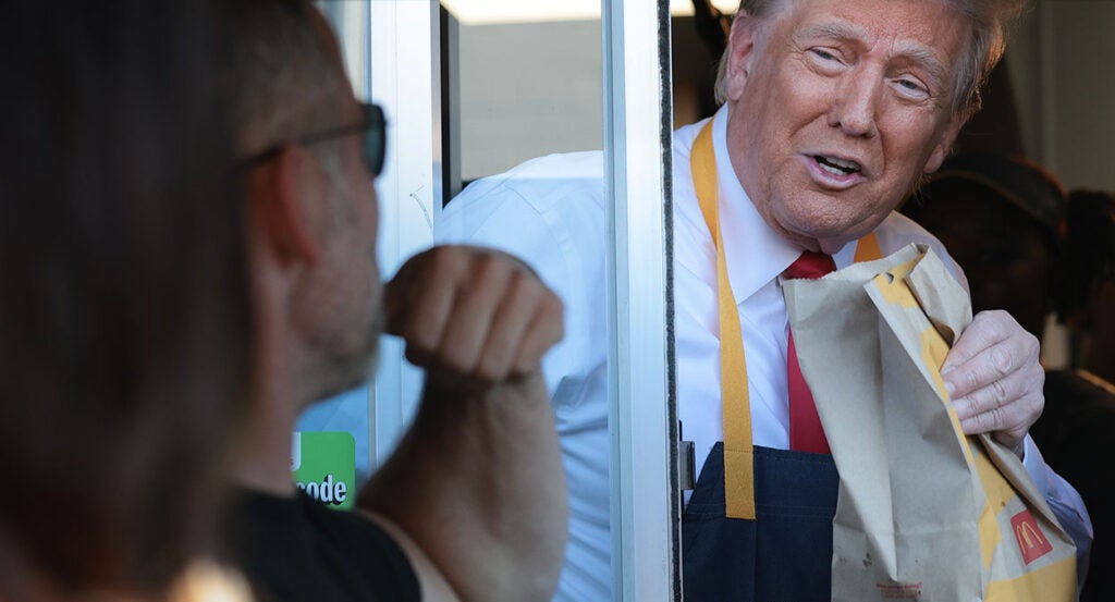 Donald Trump in a tie and McDonald's apron working a McDonald's drive-thru window, handing a bag of food to a customer