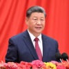 Chinese Premier Xi Jinping in a blue suit speaks in front of a red background with flowers in front of him