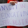 A woman holds a handwritten poster at a protest that says, "Invest in education for best ROI"