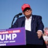 Donald Trump at an outdoor campaign rally onstage at a podium with a blue suit jacket, no tie, and a red MAGA hat