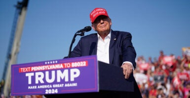 Donald Trump at an outdoor campaign rally onstage at a podium with a blue suit jacket, no tie, and a red MAGA hat